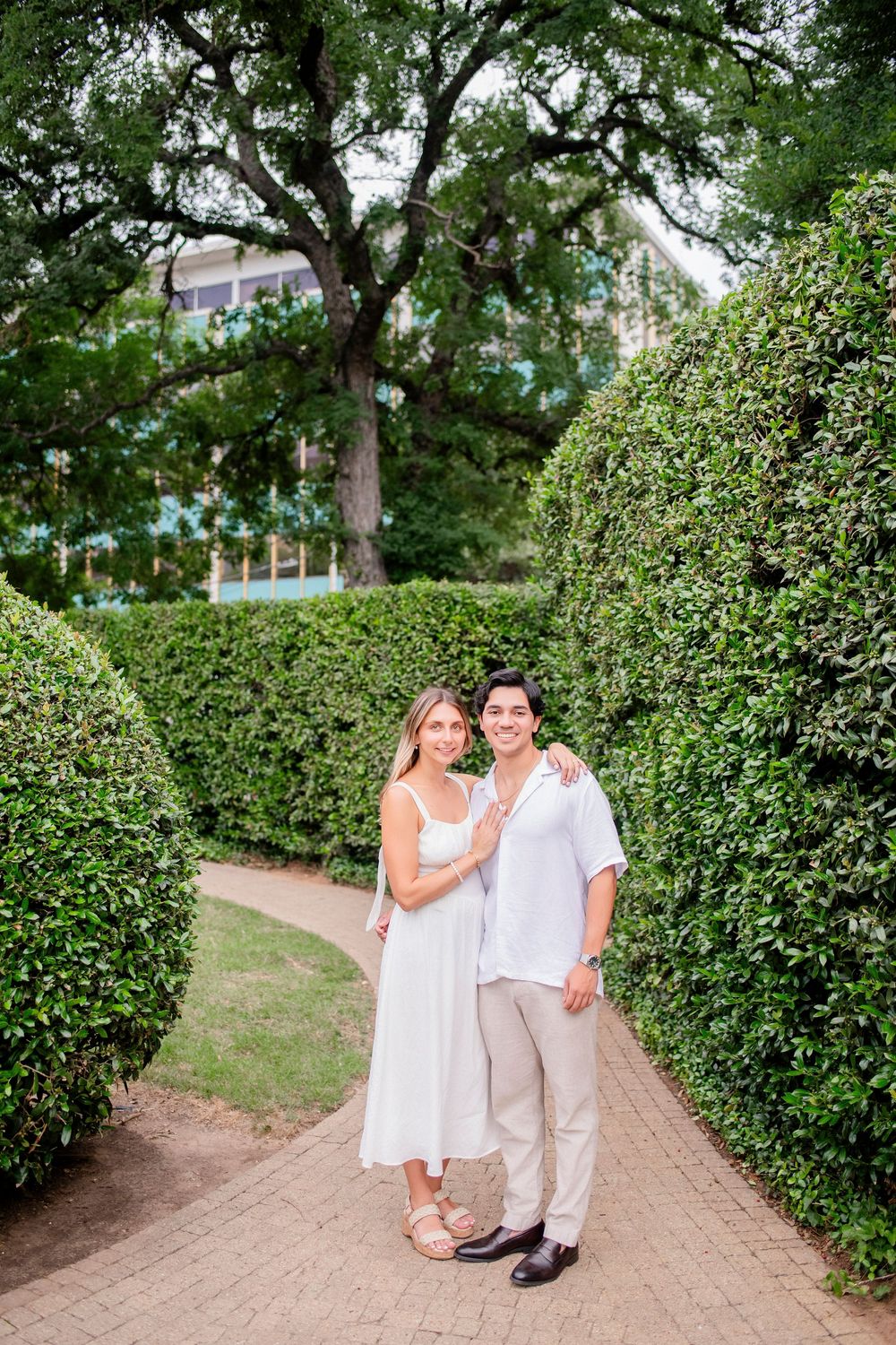 A couple in white and beige attire poses along a garden path lined with manicured hedges on a sunny day.