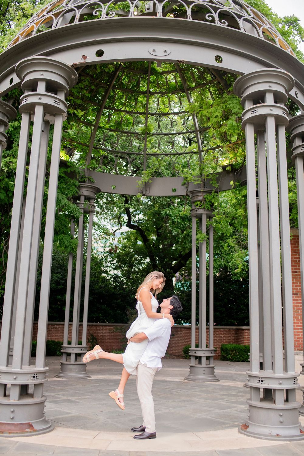 Romantic couple embraces under an ornate metal gazebo in a lush garden setting.