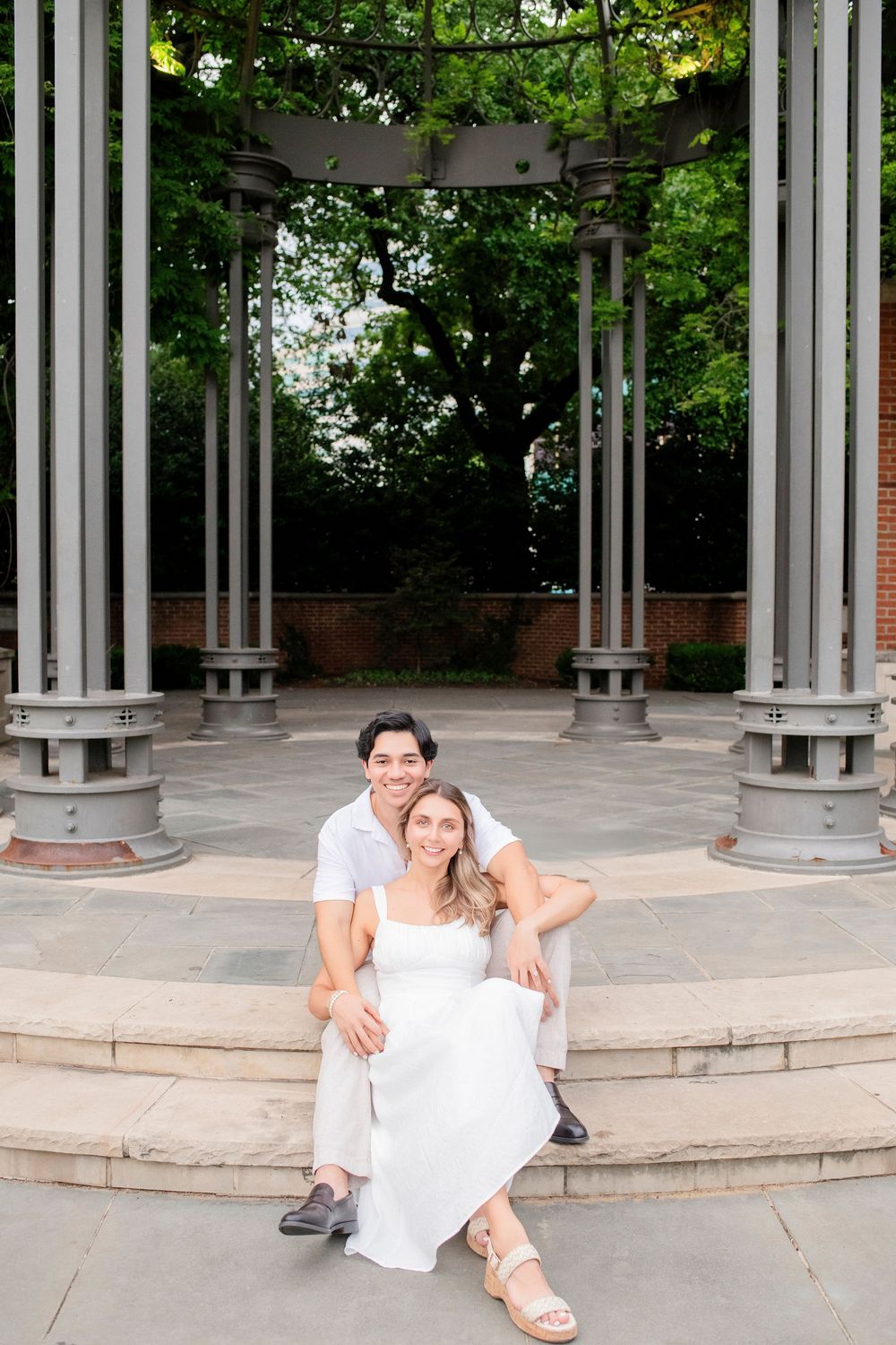 A couple sits together on stone steps under an elegant columned gazebo structure in a garden setting.