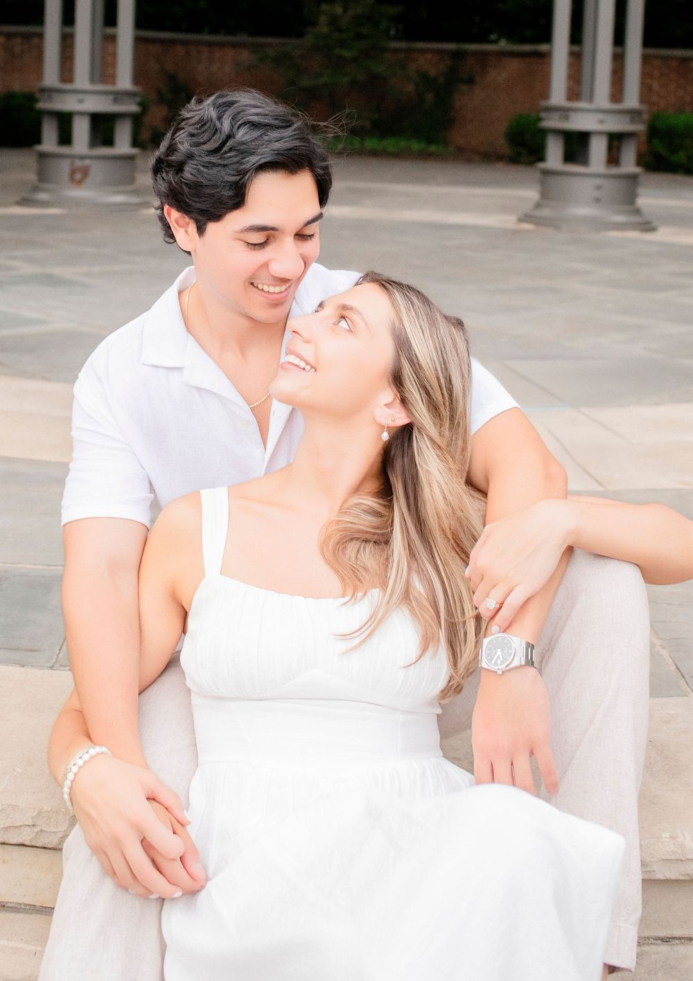 A couple in white outfits share a tender moment while sitting on stone steps outdoors.