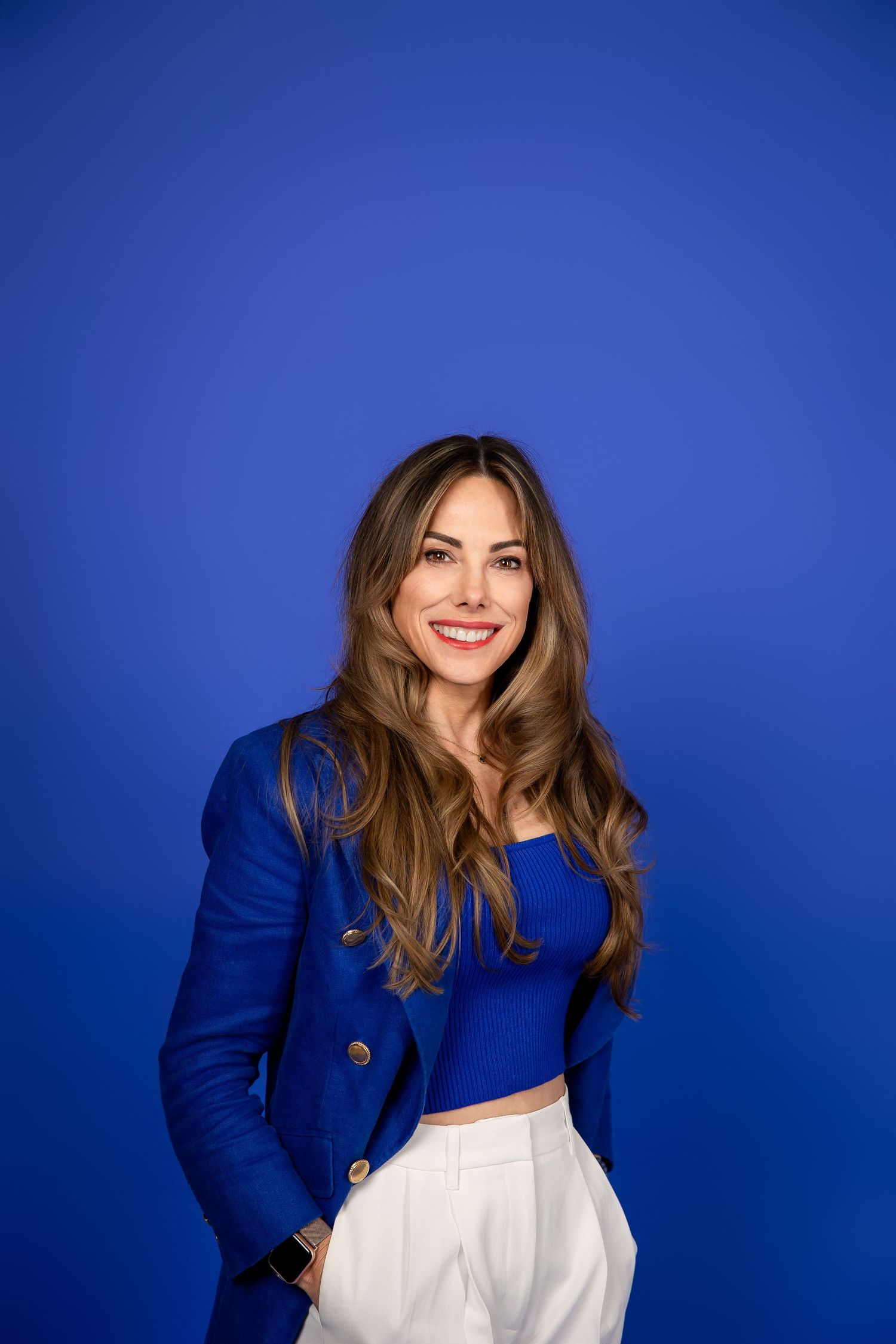 Person in a royal blue blazer and white top poses against a vibrant blue background with a warm smile.