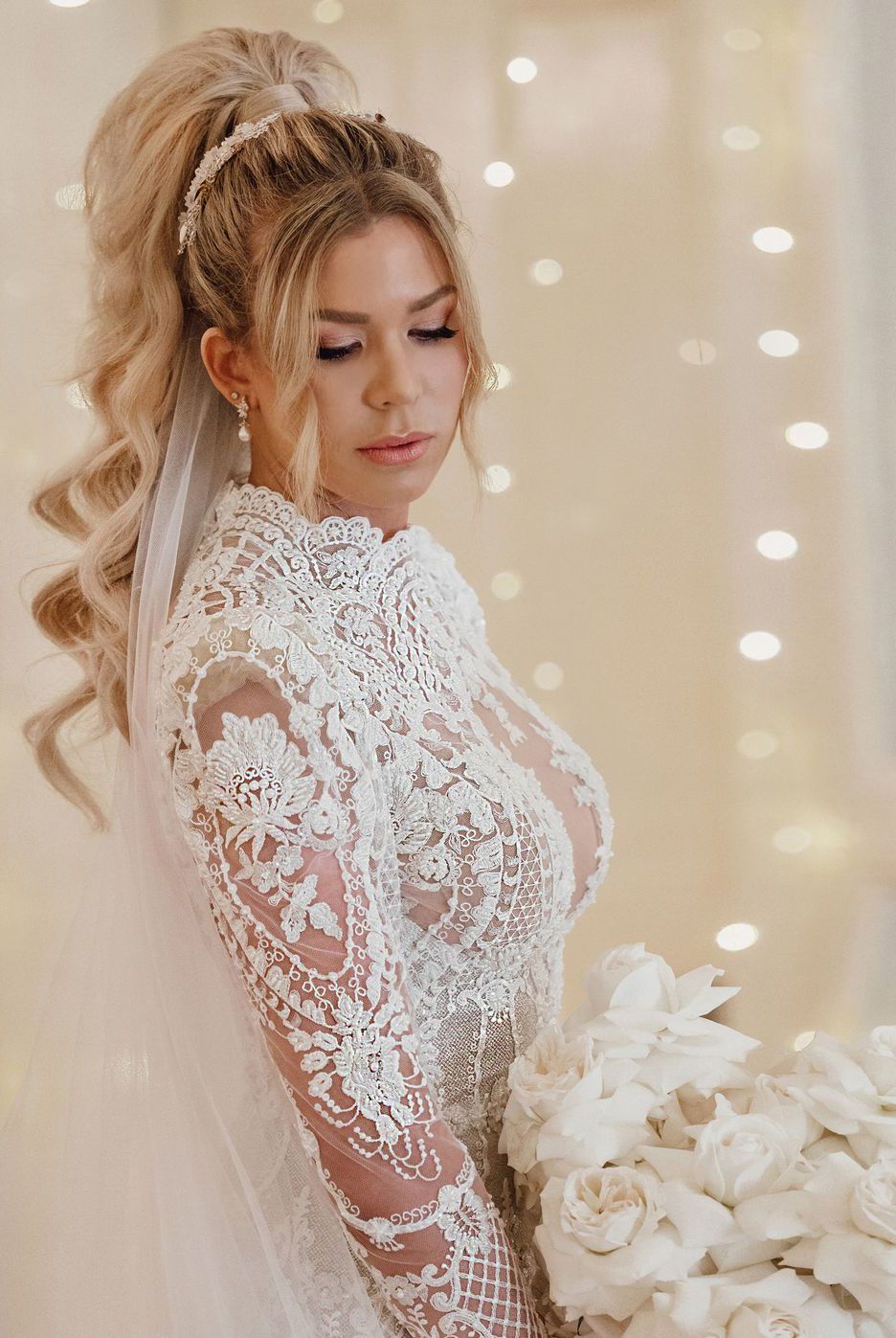 Beautiful bride in long-sleeve lace wedding gown with curly updo hairstyle holding white rose bouquet indoors.