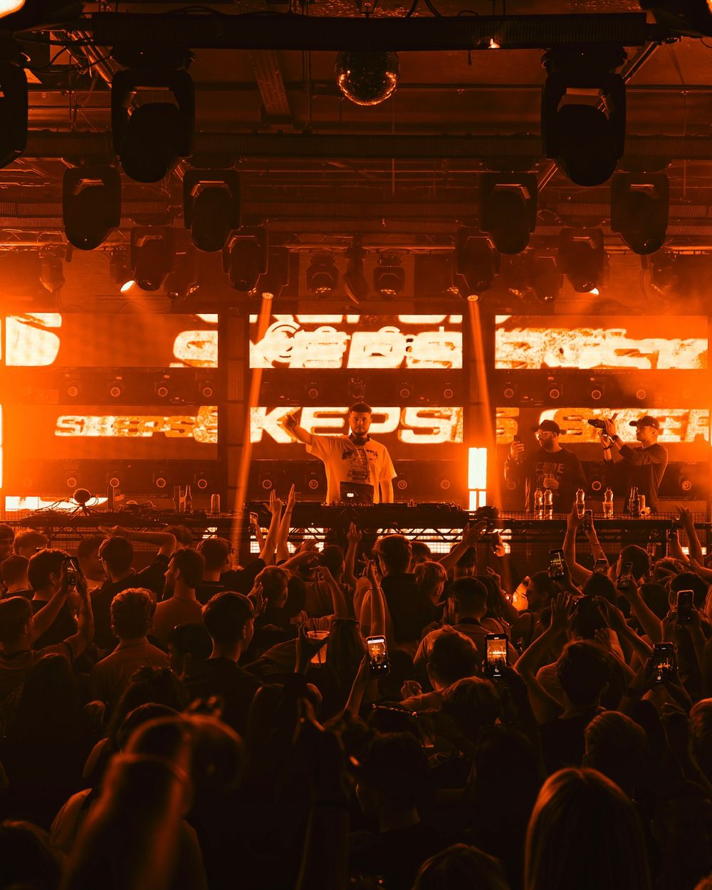 Concert crowd silhouetted against warm orange stage lighting with illuminated signage visible in the background.