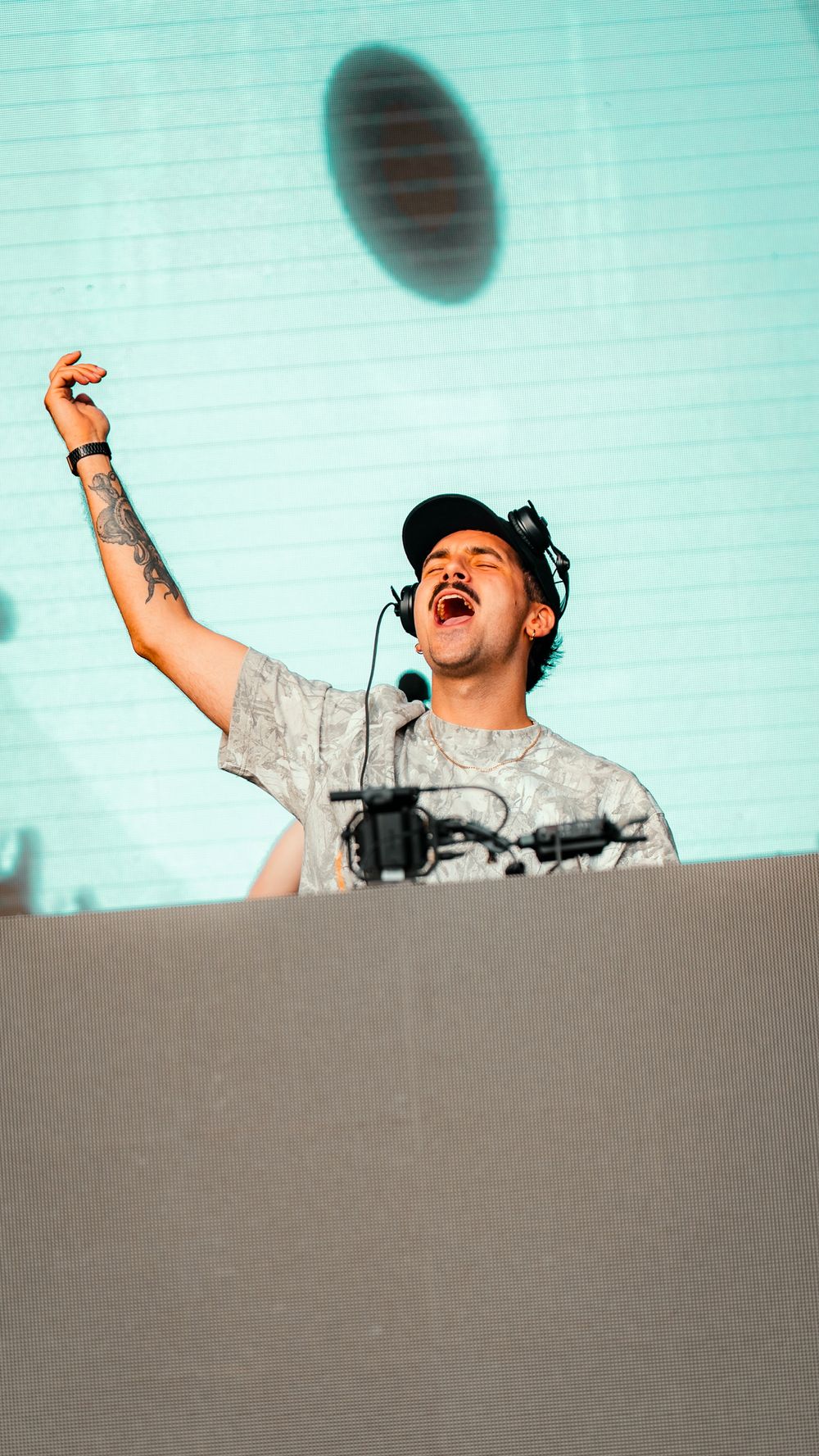 An energetic DJ performs behind decks with arms raised and headphones on against a turquoise background.