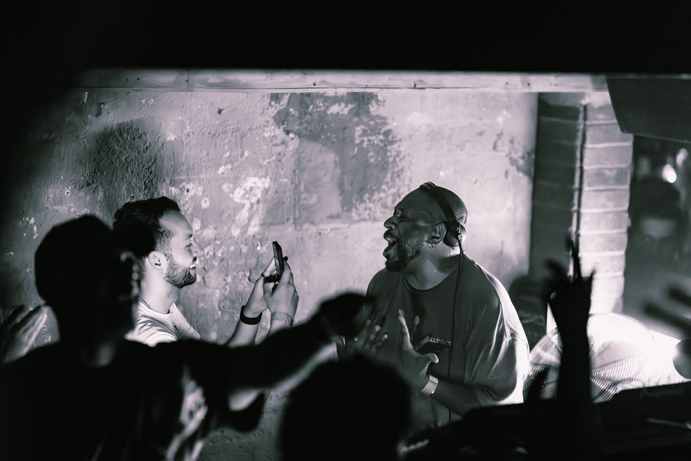 A dark atmospheric nightclub scene with silhouettes of people against a moody backlit backdrop.