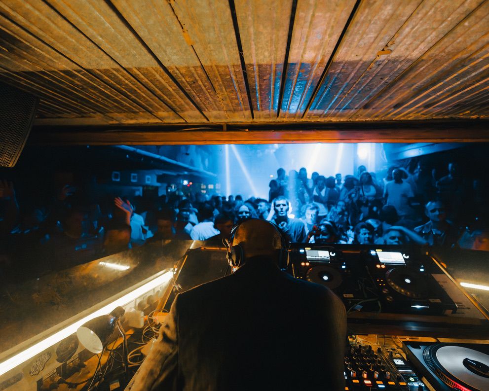 A dark nightclub scene with blue lighting shows a DJ booth overlooking a crowded dance floor.