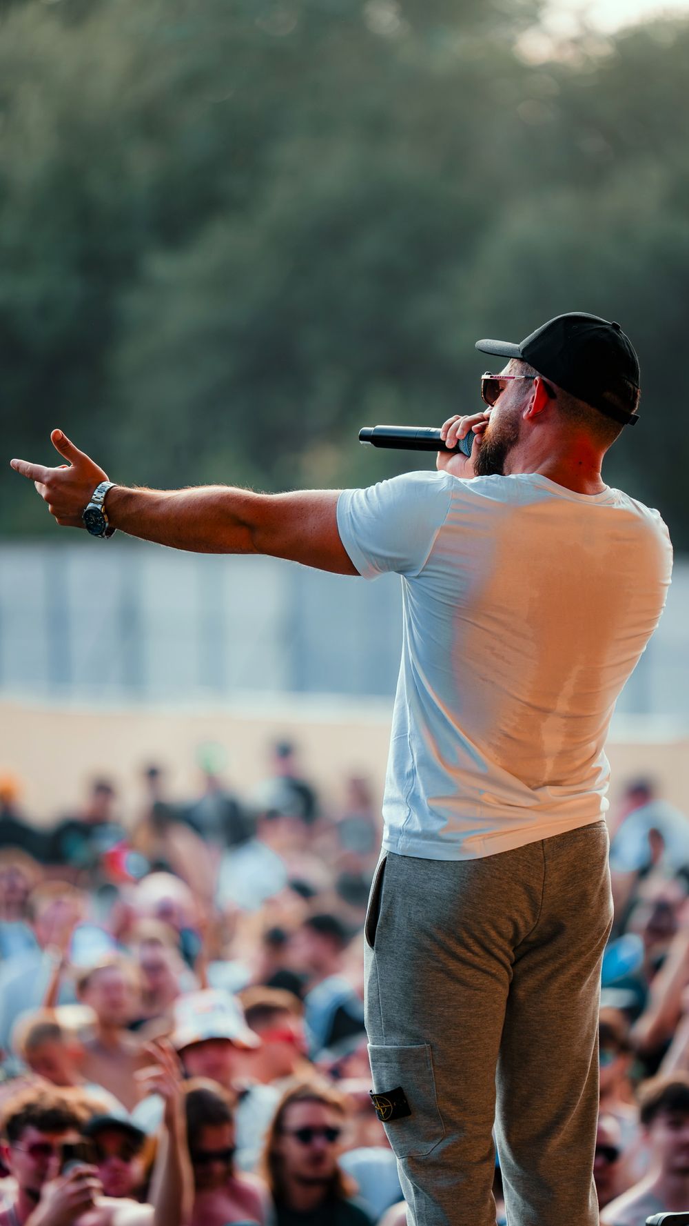 Performer in white shirt and gray pants gestures to large outdoor festival crowd from stage during daytime show.