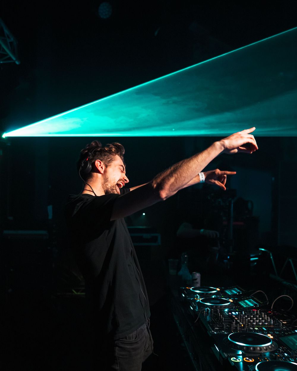 DJ performing in dark nightclub with dramatic teal lighting and raised arms.