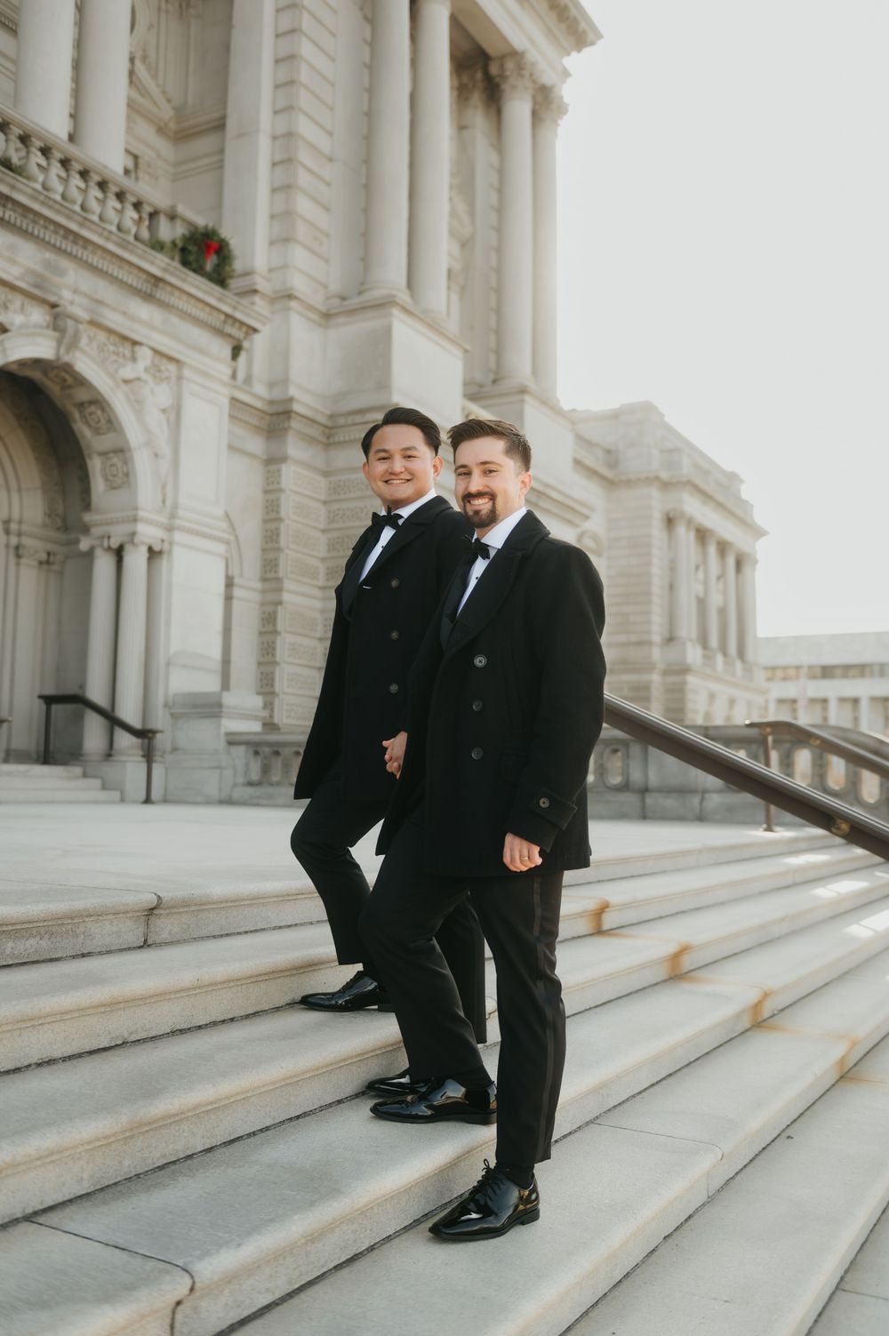 Two people in black suits sit together on elegant white marble steps of a grand building.