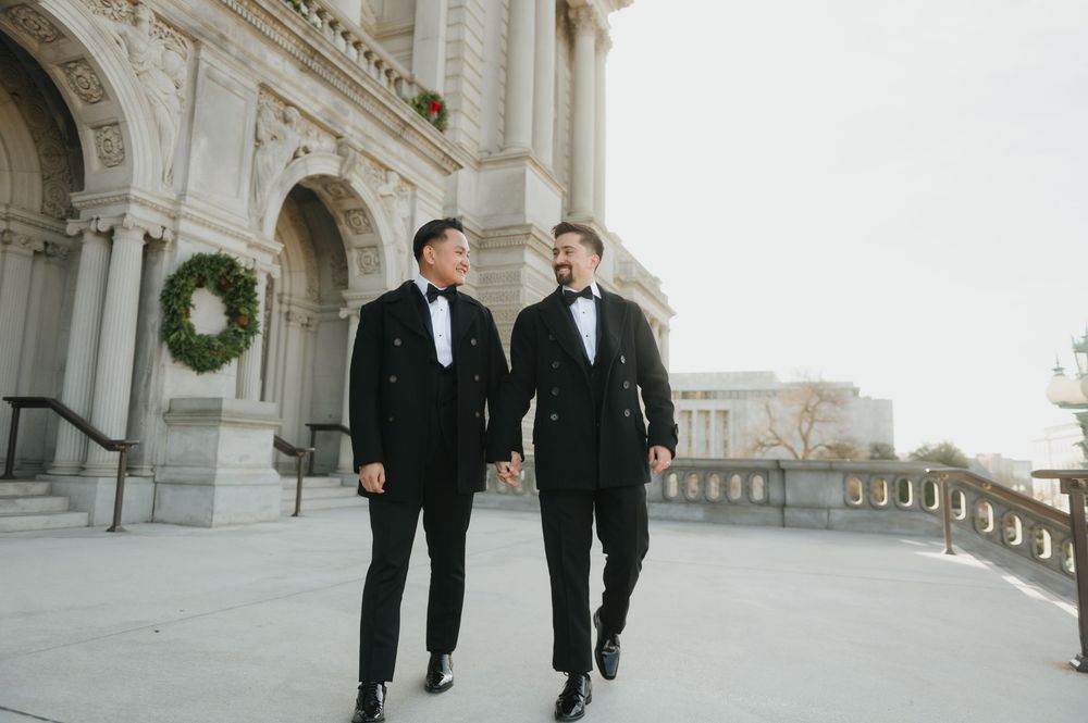 Two people in black suits walk and smile together along an elegant architectural corridor with stone columns.
