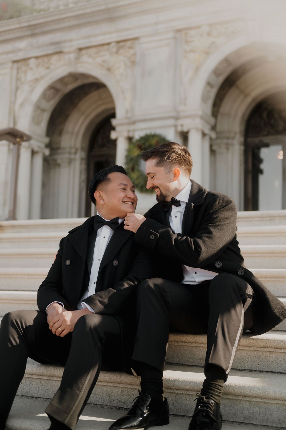 Two people in tuxedos share a candid moment while sitting on stone steps of an elegant building.