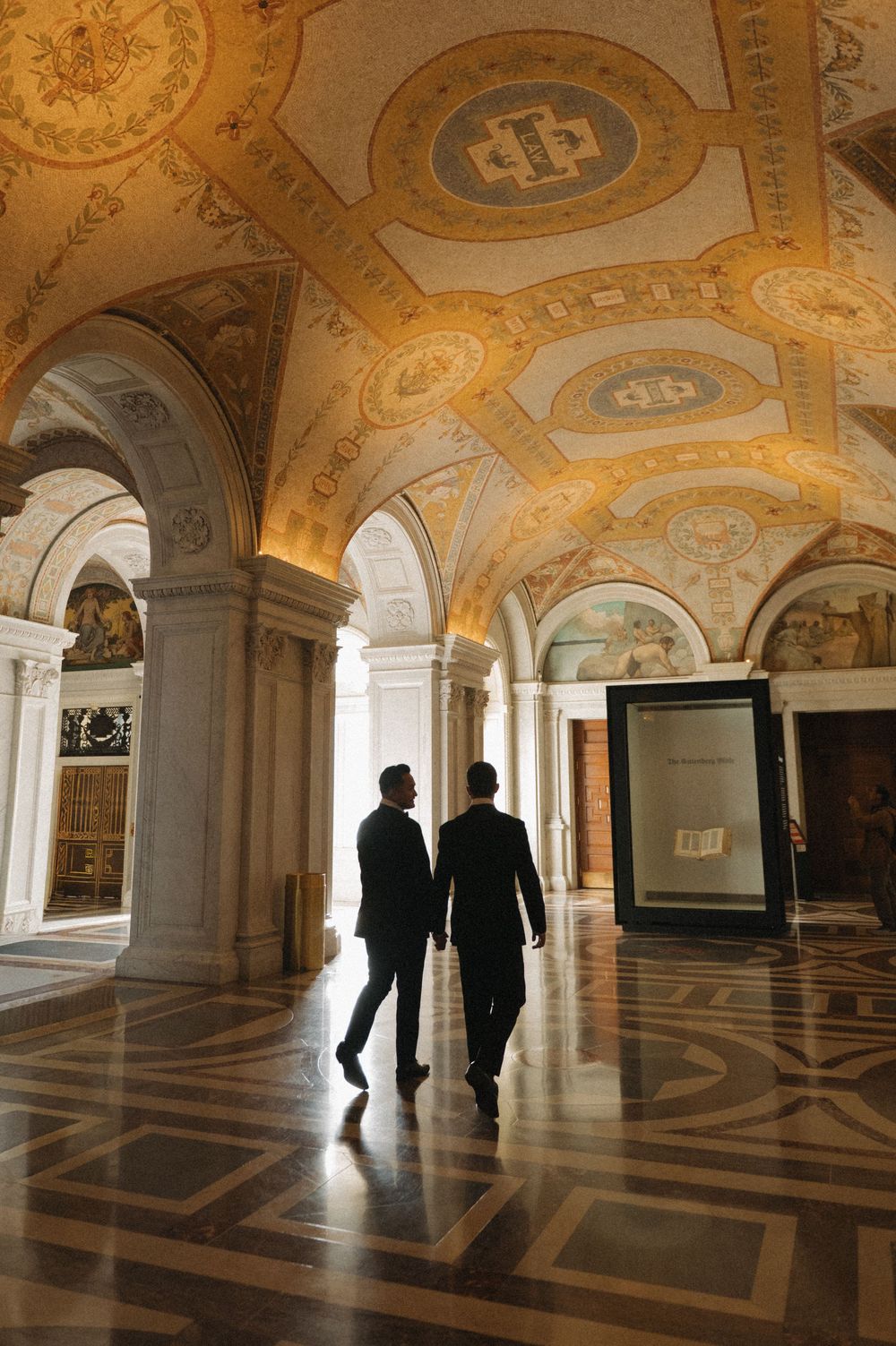 Two silhouettes walk through an ornate hallway with gilded ceilings and patterned marble floors.