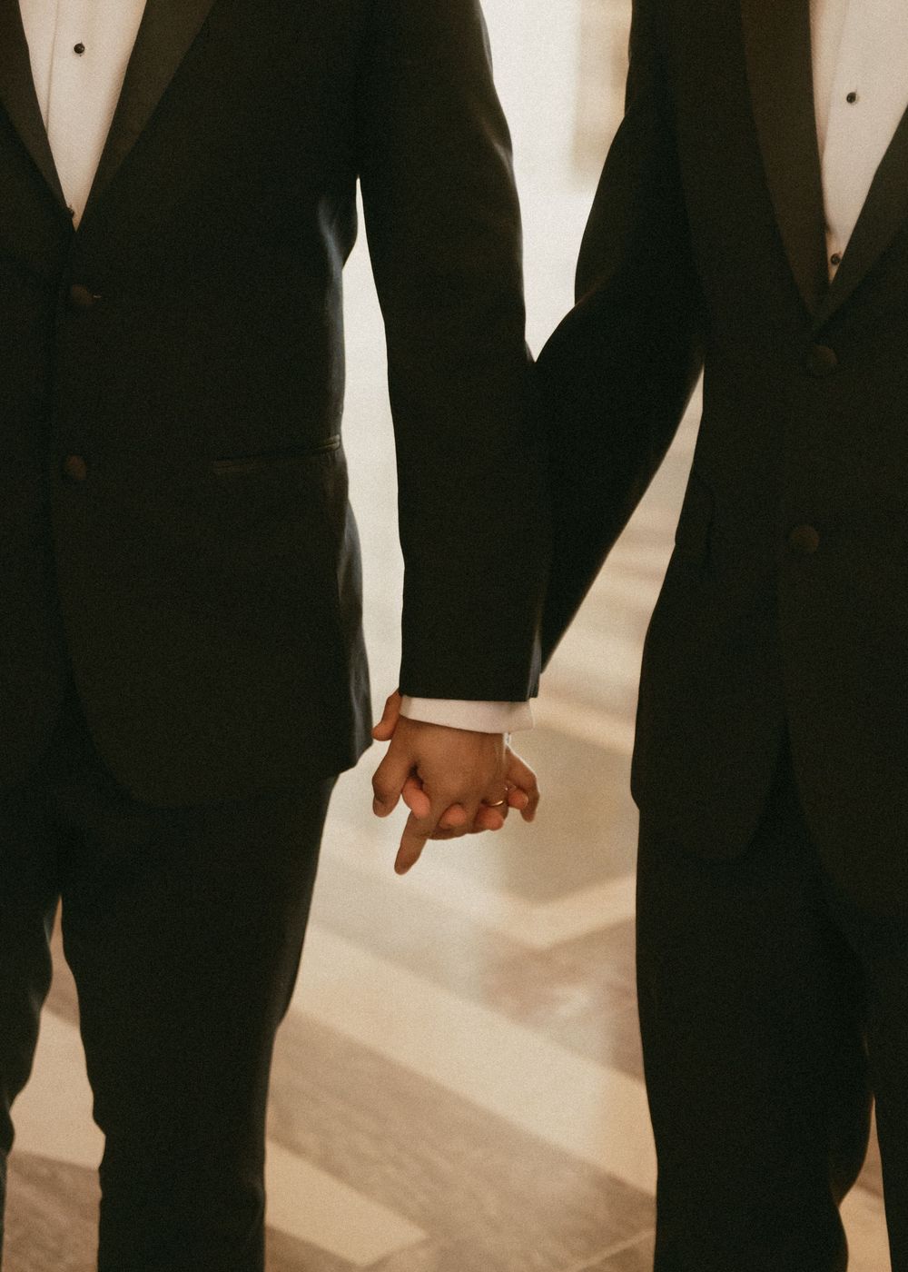 Two people in black tuxedos holding hands in an intimate wedding moment against a light background.