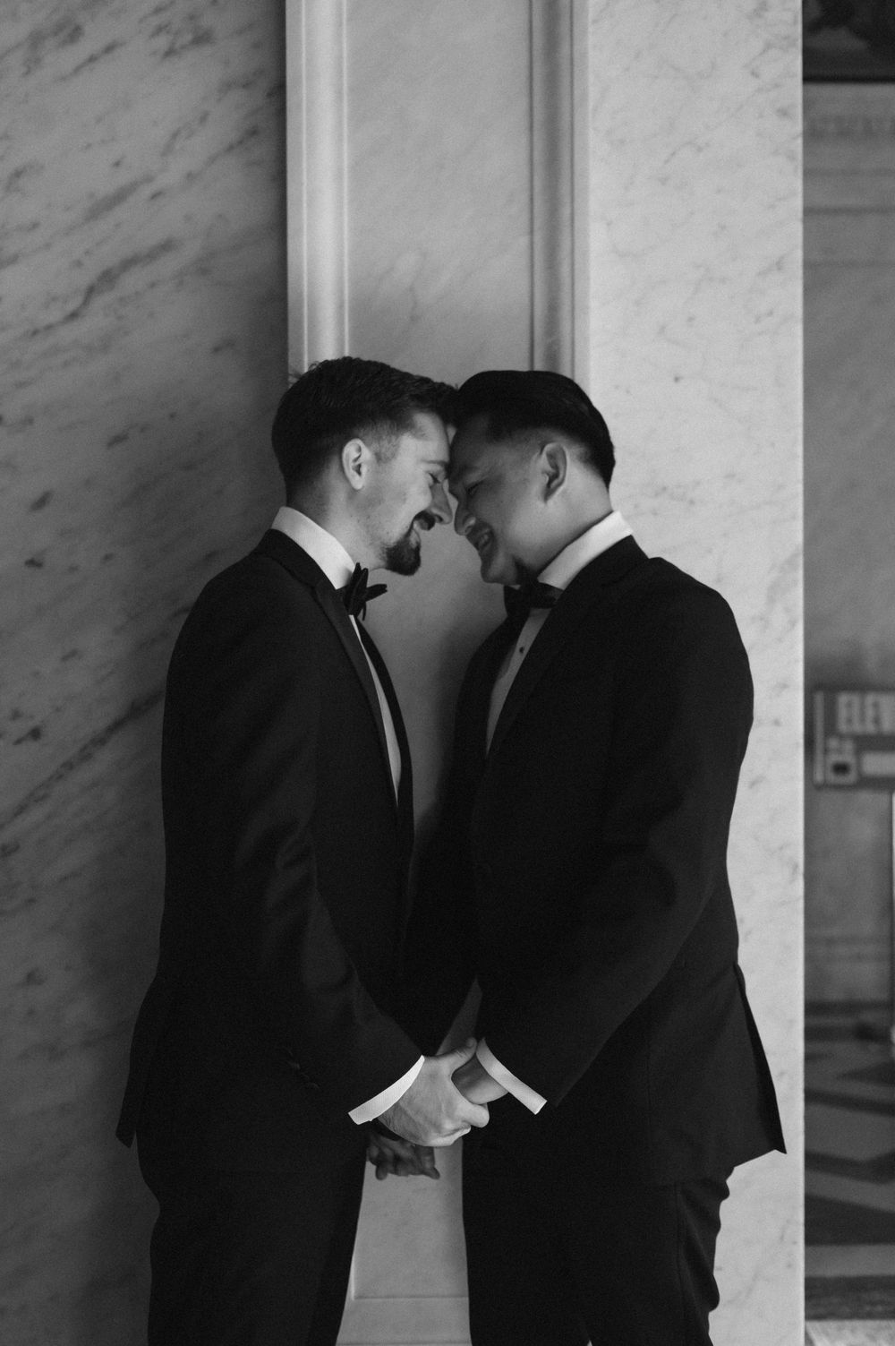 A romantic black and white photo of a couple in tuxedos sharing an intimate moment against a marble column.