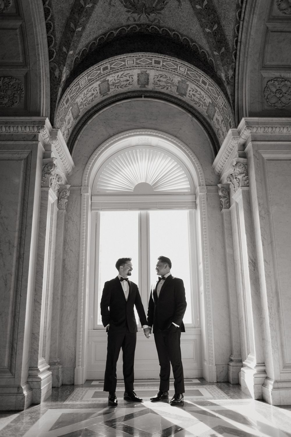 Two figures in formal attire stand in conversation within an ornate architectural corridor with arched windows.