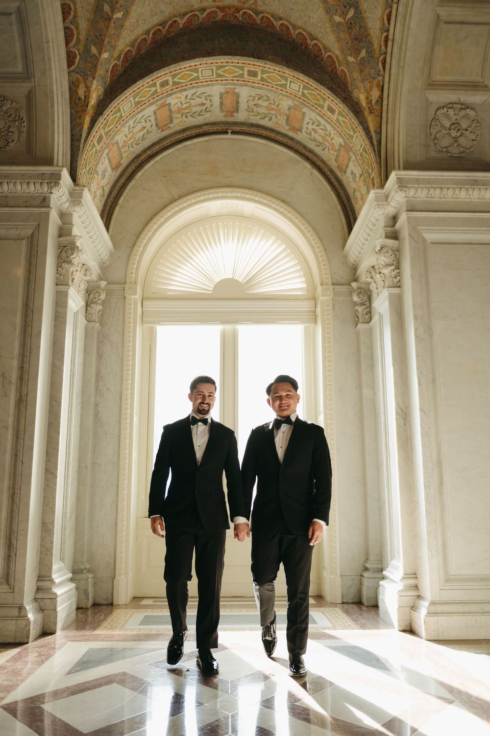 Two people in black tuxedos holding hands while walking through an elegant marble hallway with arched ceilings.