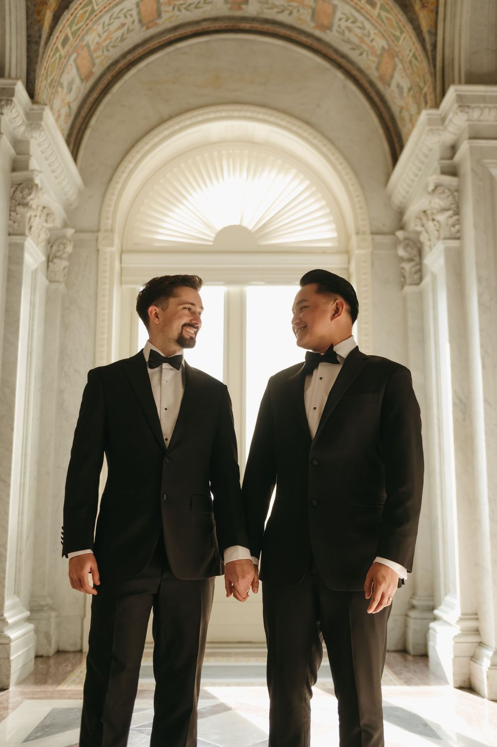 Two grooms in black tuxedos holding hands and smiling at each other under an elegant arched hallway.