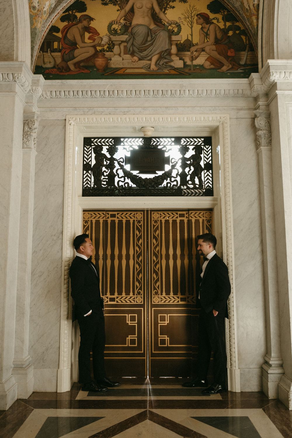 Two figures in tuxedos stand in an elegant marble hallway with ornate gold gates and decorative ceiling murals.