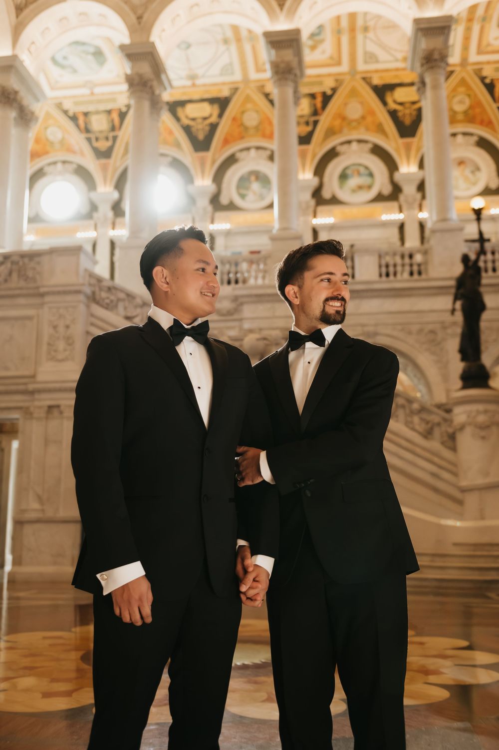 A couple in formal black tuxedos standing together in an ornate historic building with decorated ceilings.