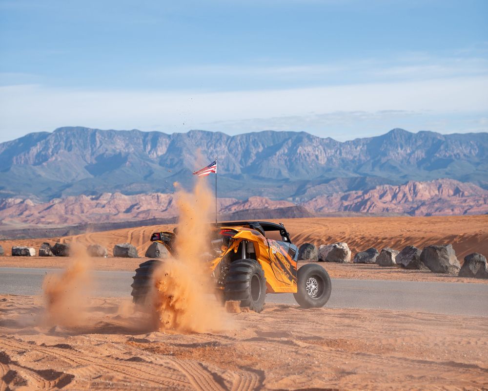 Yellow off-road buggy racing through desert sand dunes with mountains in background kicking up dust cloud.