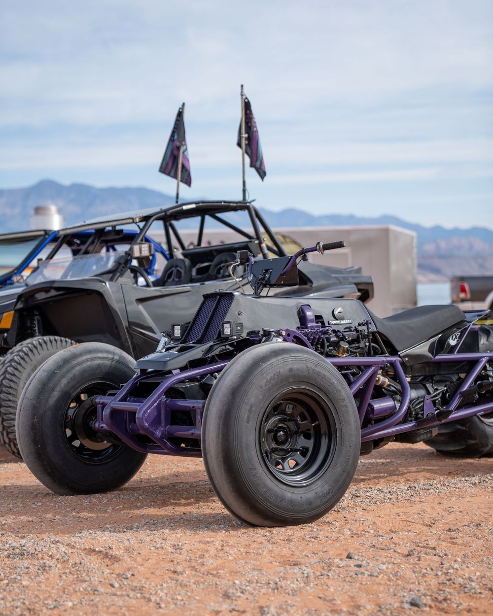 Custom off-road dune buggy with purple frame and large tires parked on desert terrain with mountains in background.