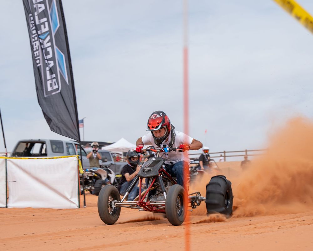 ATV rider racing on red dirt track kicking up dust at an off-road motorsport event with banners visible.