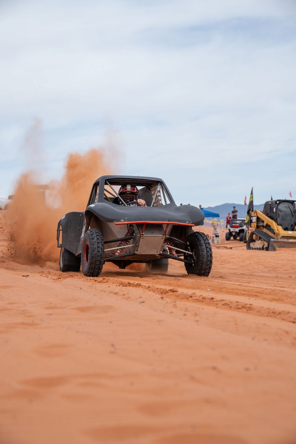 A black off-road buggy kicks up red dirt clouds while racing across a sandy desert track under a cloudy sky.