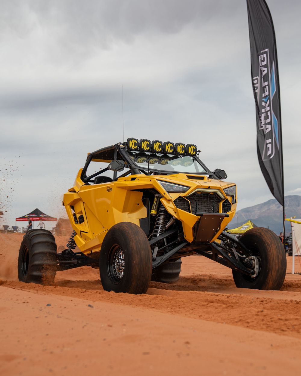 Yellow off-road UTV racing on red dirt terrain at a desert event, with a branded banner flag and cloudy skies in the background.