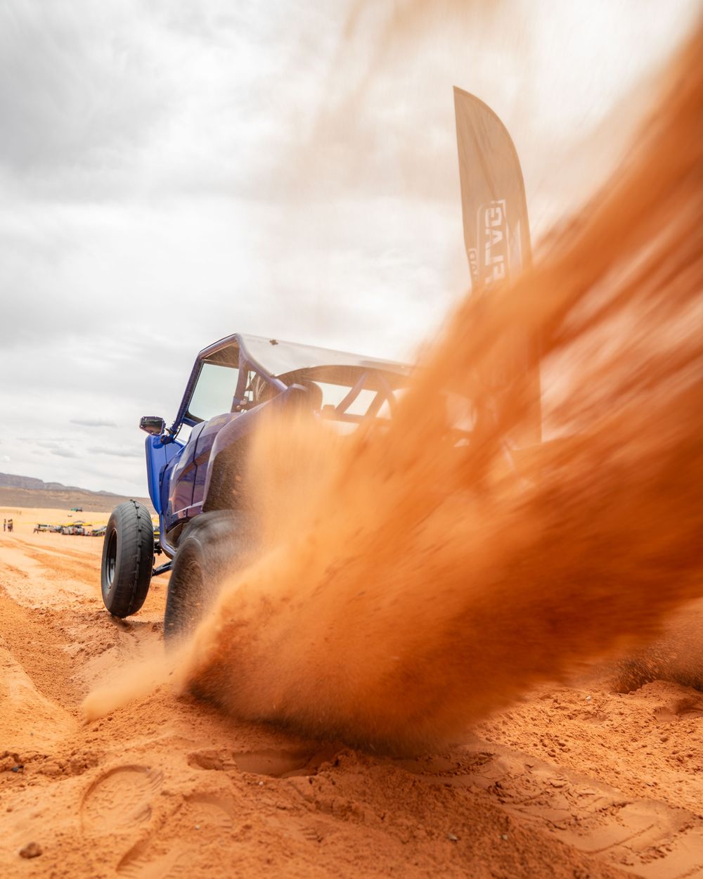 Blue off-road buggy kicking up dramatic red sand dust cloud in desert terrain during rally race.