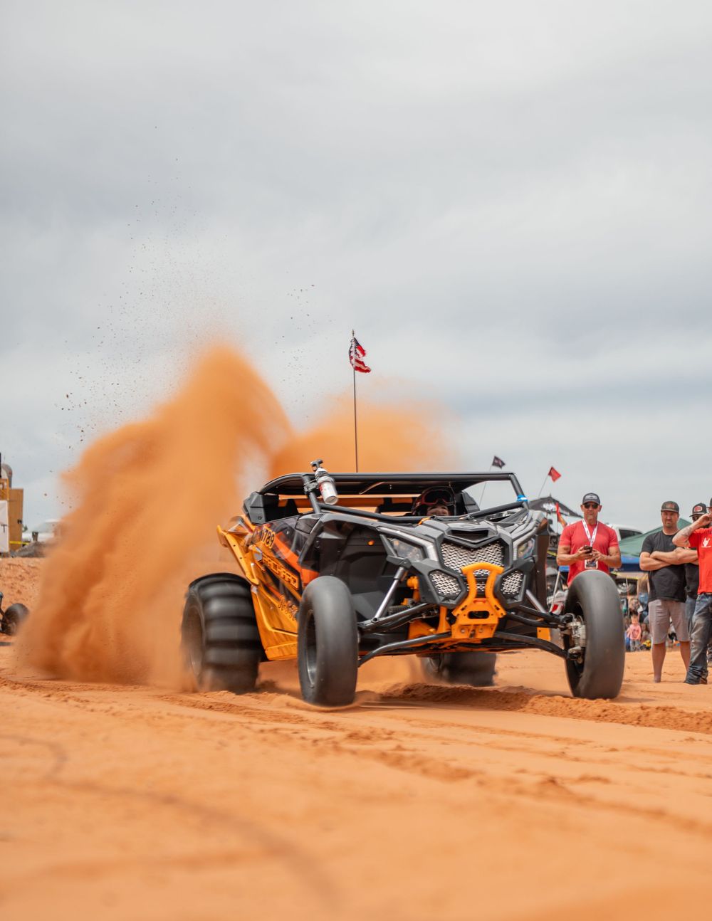 Orange off-road UTV kicking up red dirt sand while racing through a desert terrain at an outdoor motorsport event.