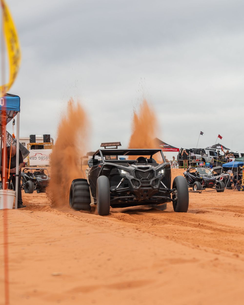 Black off-road UTV kicking up dramatic sand plumes on a red dirt track at an outdoor motorsport event.