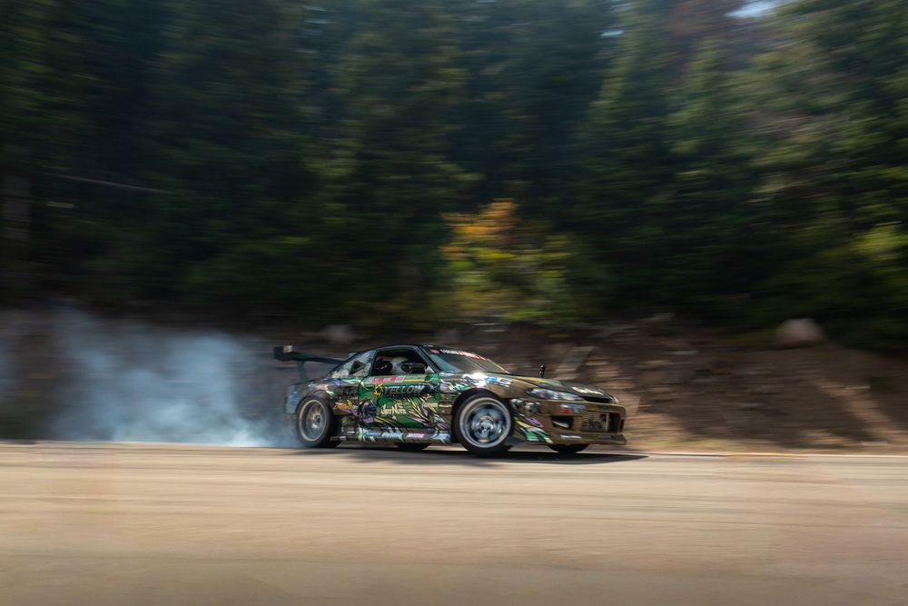 A high-performance race car drifts around a forested track with smoke billowing from its tires.