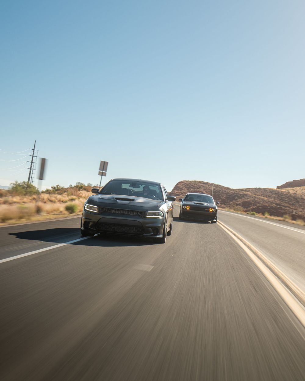 Two Dodge Chargers racing on a desert highway under a clear blue sky with rocky hills in the background.