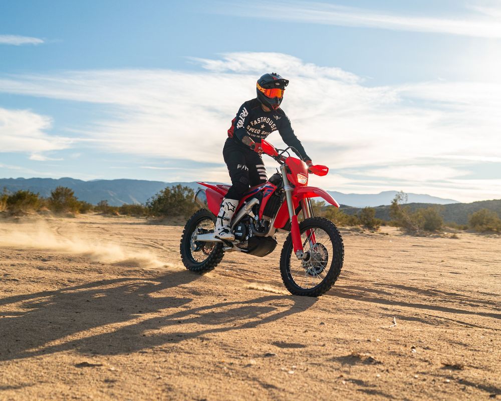 A motocross rider performs a wheelie on a red dirt bike across a sandy desert landscape with mountains in the background.