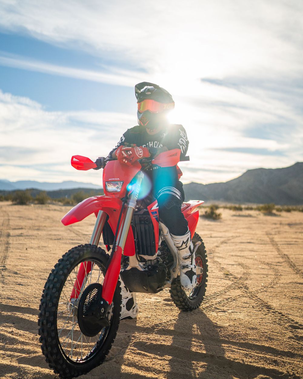 Motocross rider on red dirt bike in desert landscape with mountains and dramatic sky backdrop.