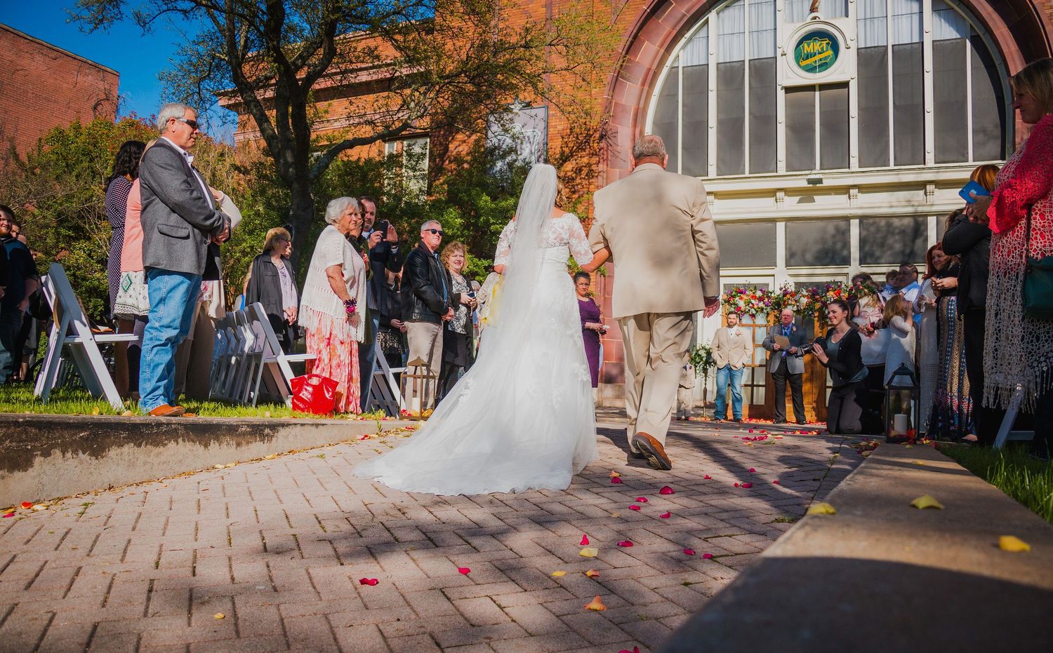 Bride Misty walks with her father to her Groom Larry at the Katy Depot entrance and floral altar in Denison, TX. by Wolfpack Wedding Co.