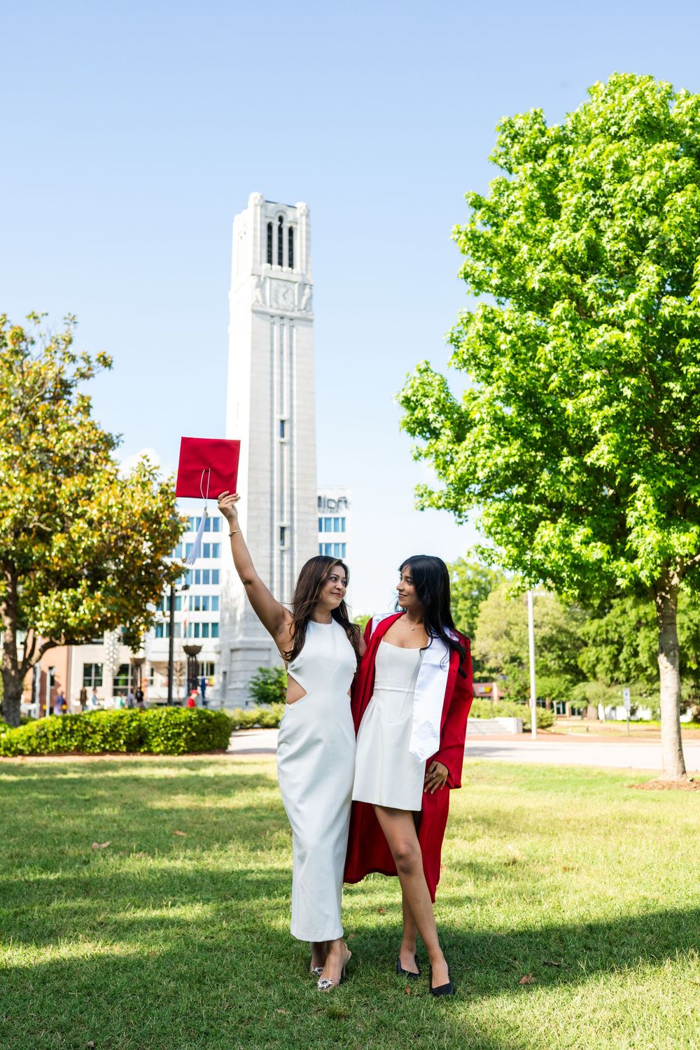Graduates celebrate with diplomas on campus lawn with iconic university tower in background.