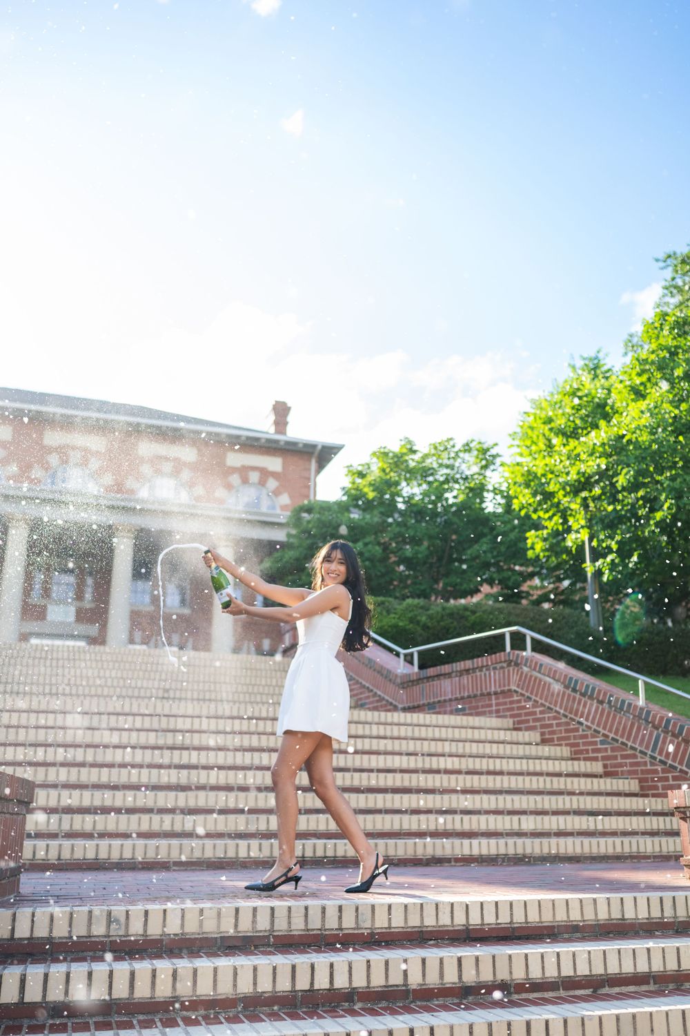 Graduate poses joyfully on stone steps during graduation photoshoot on sunny day.