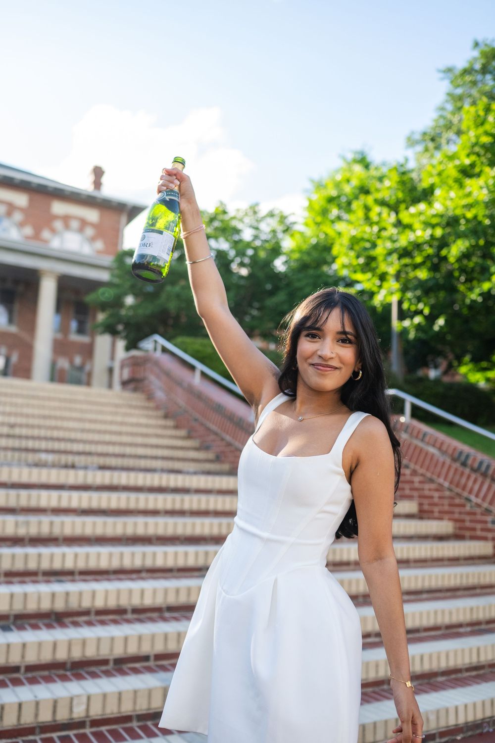 A person in a flowing white dress poses joyfully on outdoor stone steps surrounded by greenery.