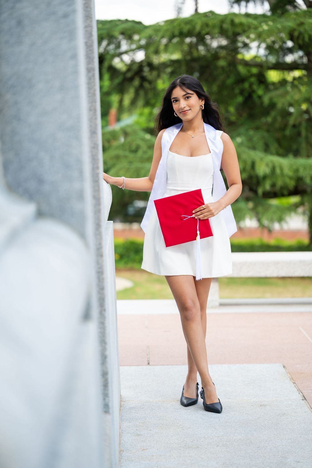 Woman in white dress and heels poses elegantly between concrete pillars while holding a red clutch purse.