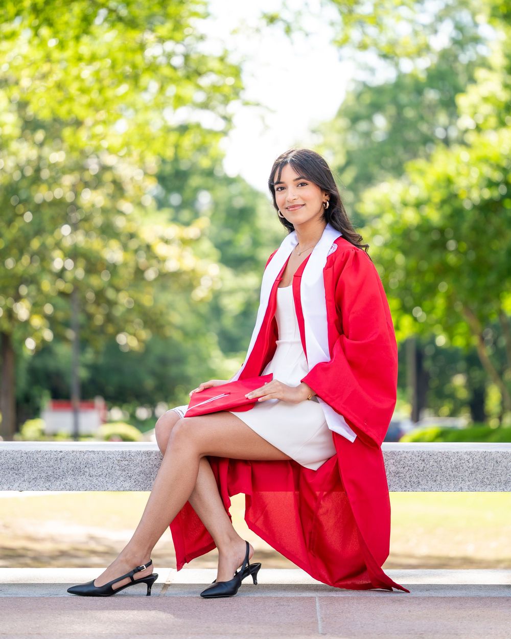 Graduate in red and white dress poses outdoors on stone wall during sunny summer photoshoot.