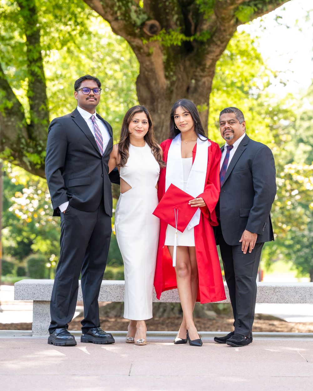 Four people in formal attire standing together on stone steps in a park with lush green trees in the background.