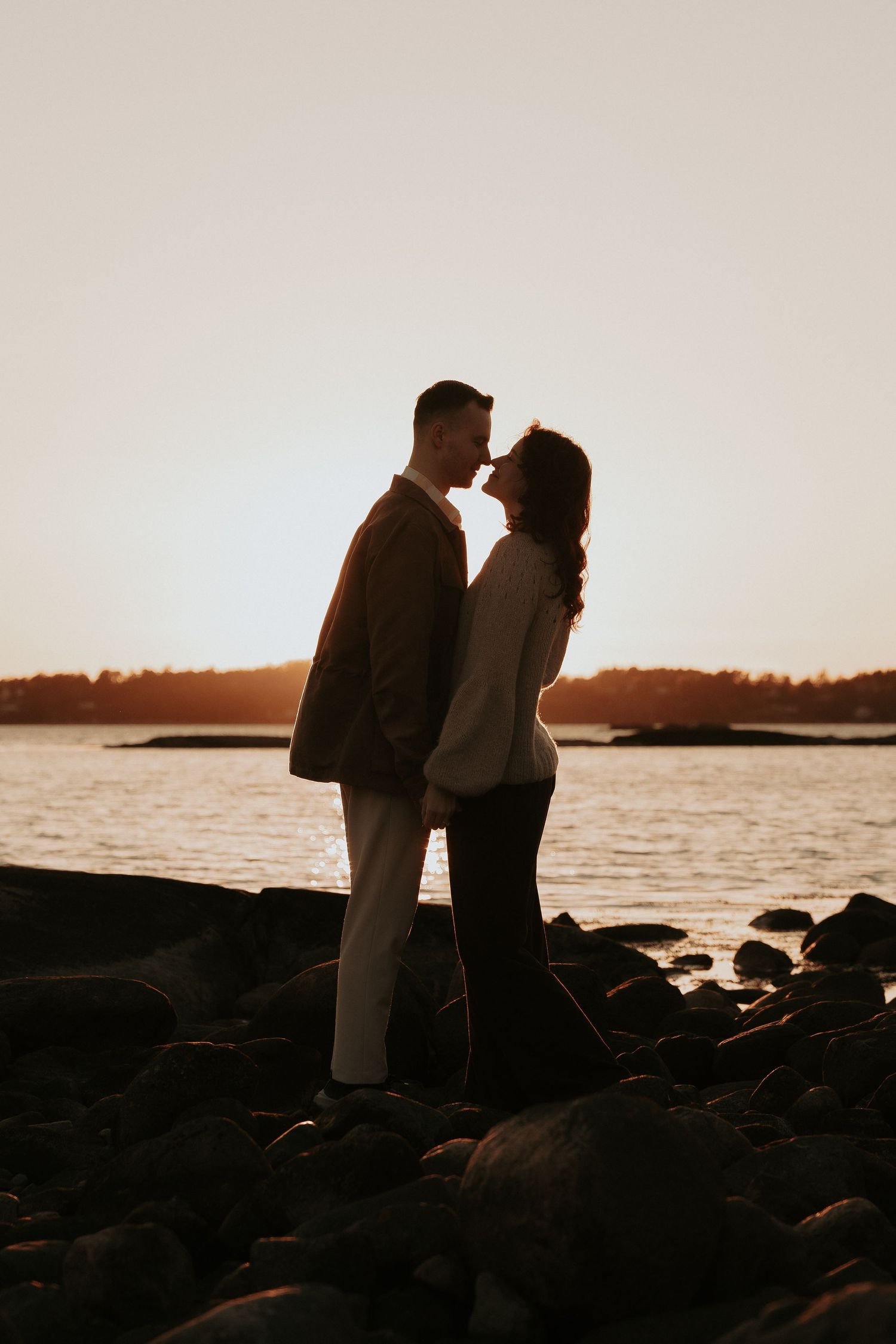 Silhouettes of a couple standing on rocky shore during sunset by the ocean.