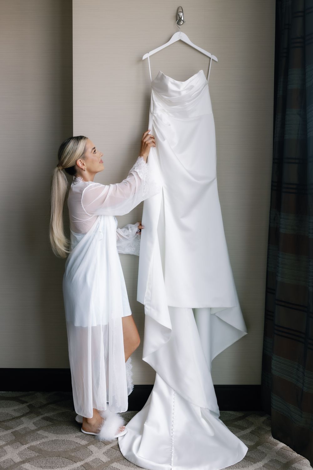 Bride in white robe admiring her elegant wedding dress hanging on display.