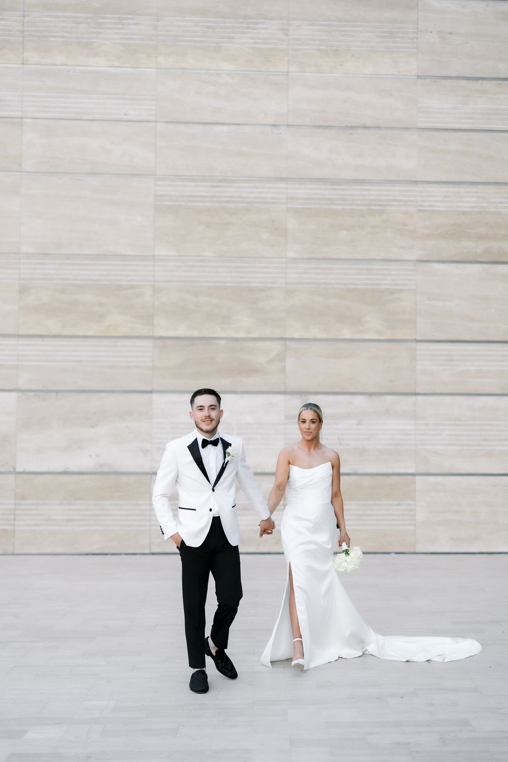 A wedding couple sits playfully on light stone steps in formal attire with a flowing white dress.