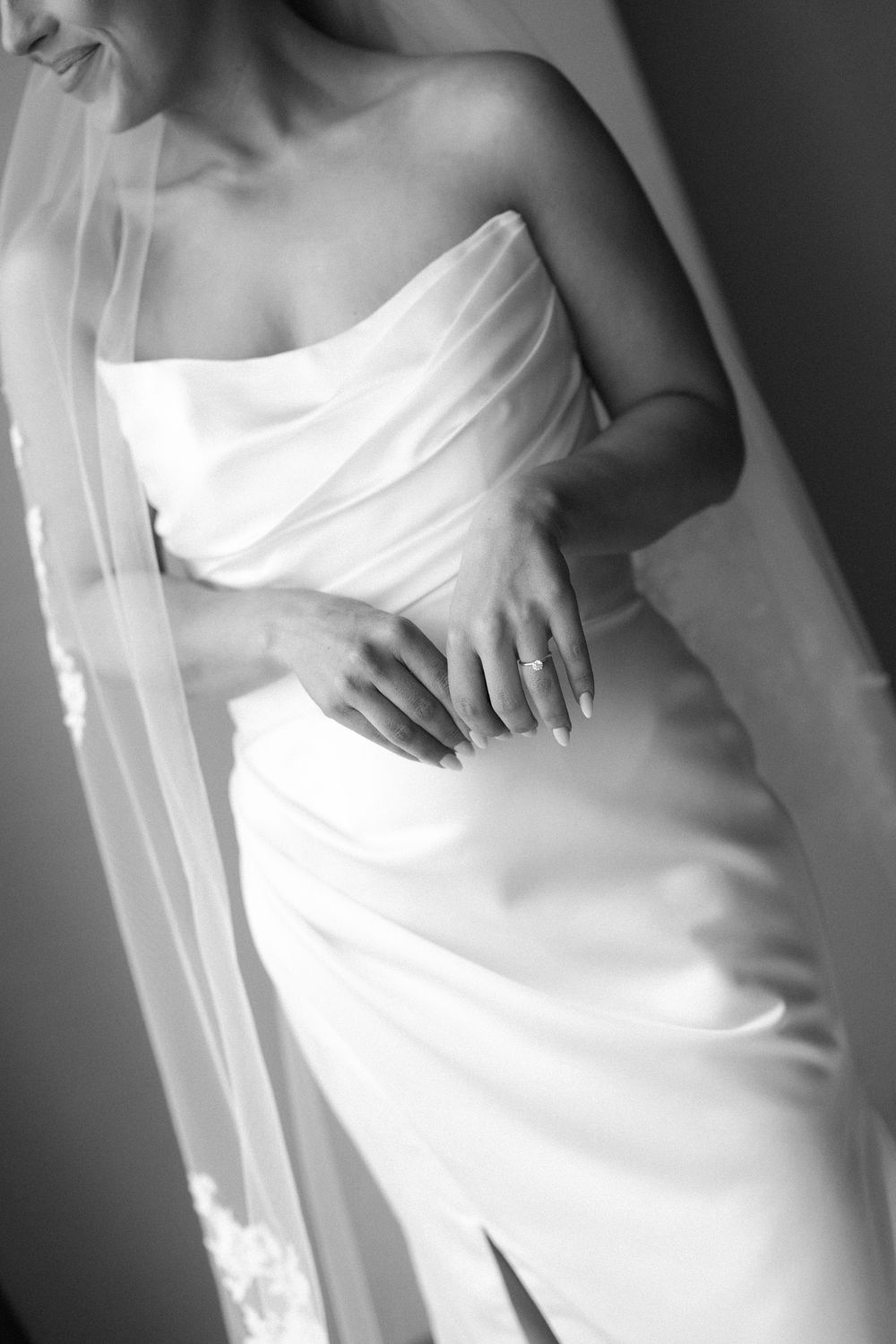 Black and white photo of a bride in a strapless wedding gown with veil, hands folded showing engagement ring.