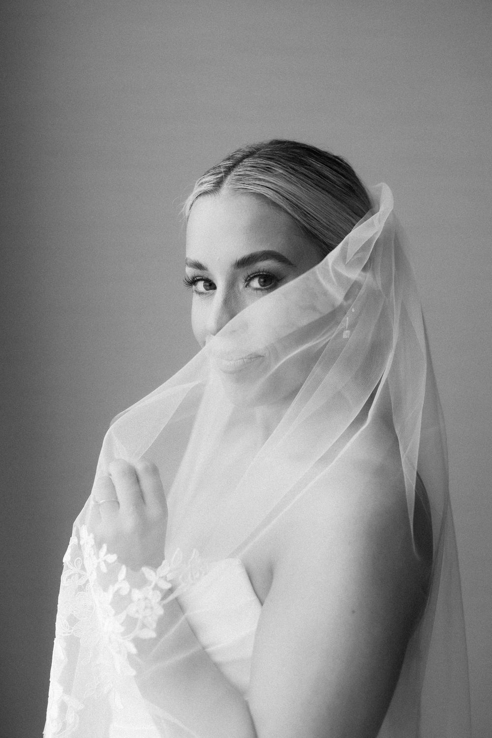 Black and white bridal portrait of a blonde woman with a lace veil draped over her face, gazing softly at the camera.