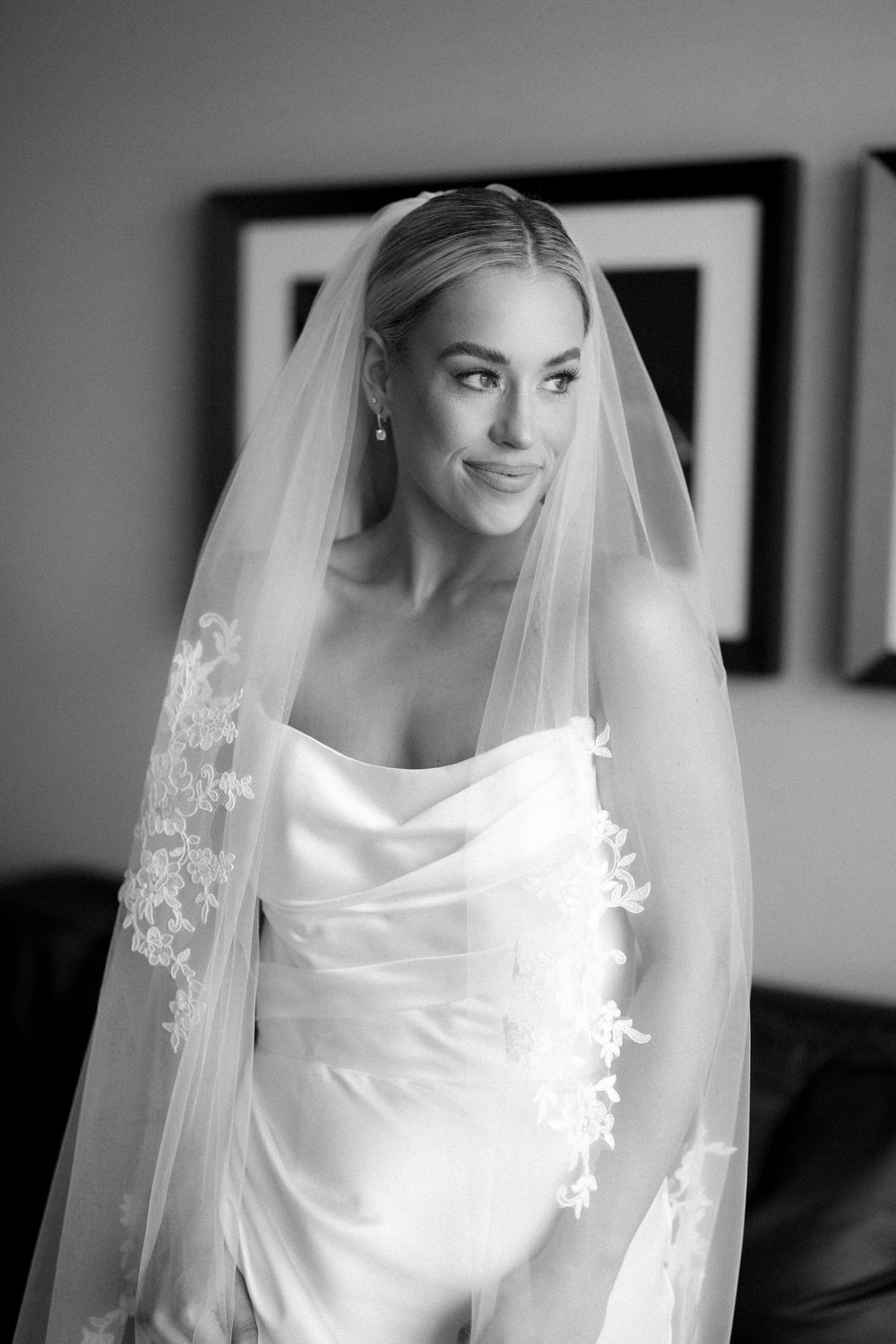 Black and white portrait of a bride in a strapless gown and lace-trimmed veil, smiling softly indoors.
