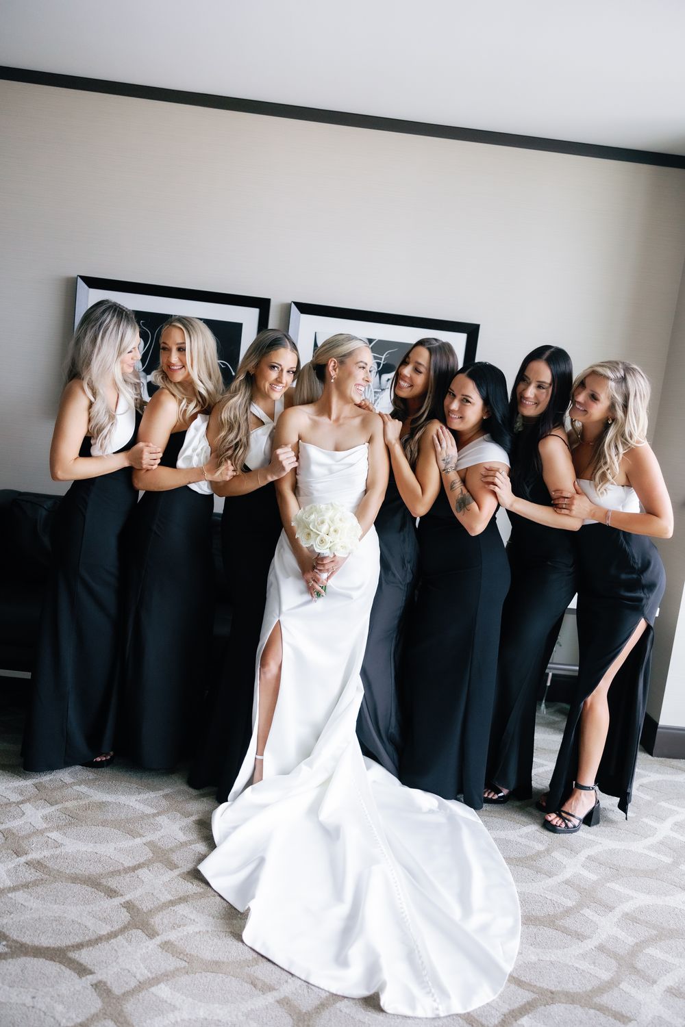 Wedding party poses playfully with bride in white gown surrounded by bridesmaids in black dresses.