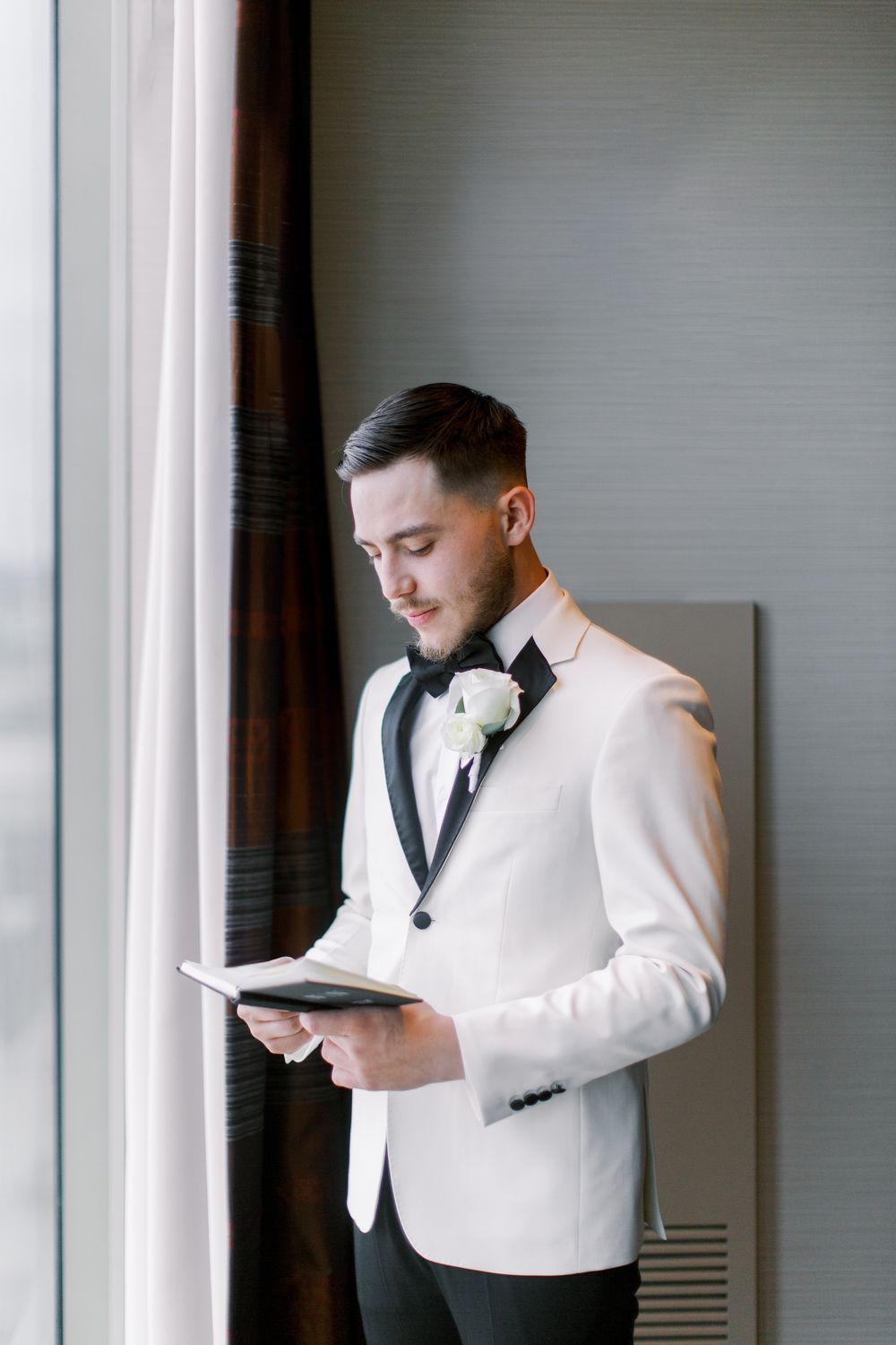 A groom in formal attire checks his phone while standing by large windows.